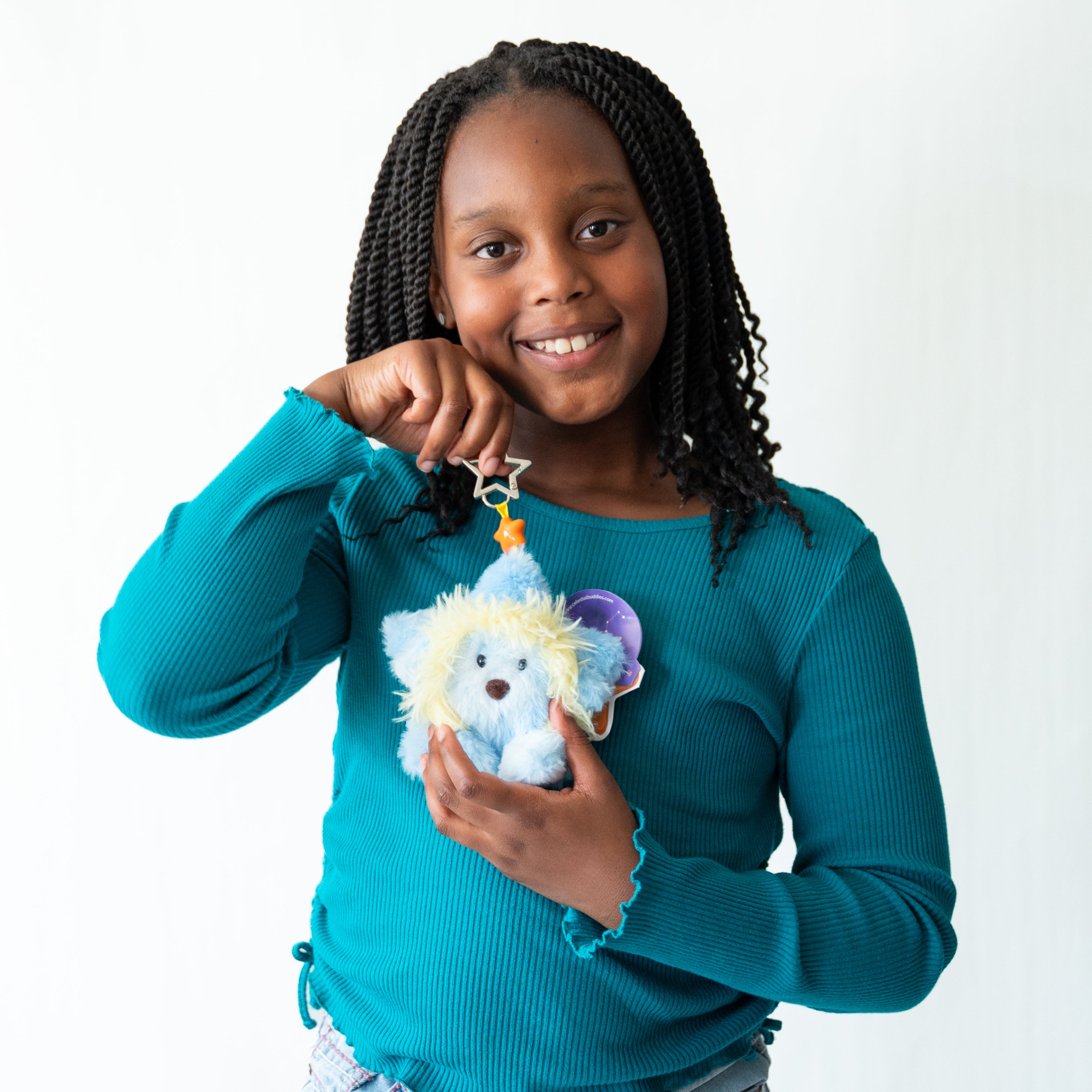 A young girl with braided hair smiles while holding a plush star-shaped keychain featuring a blue Lion with yellow hair . She is wearing a teal long-sleeve top against a plain white background.
