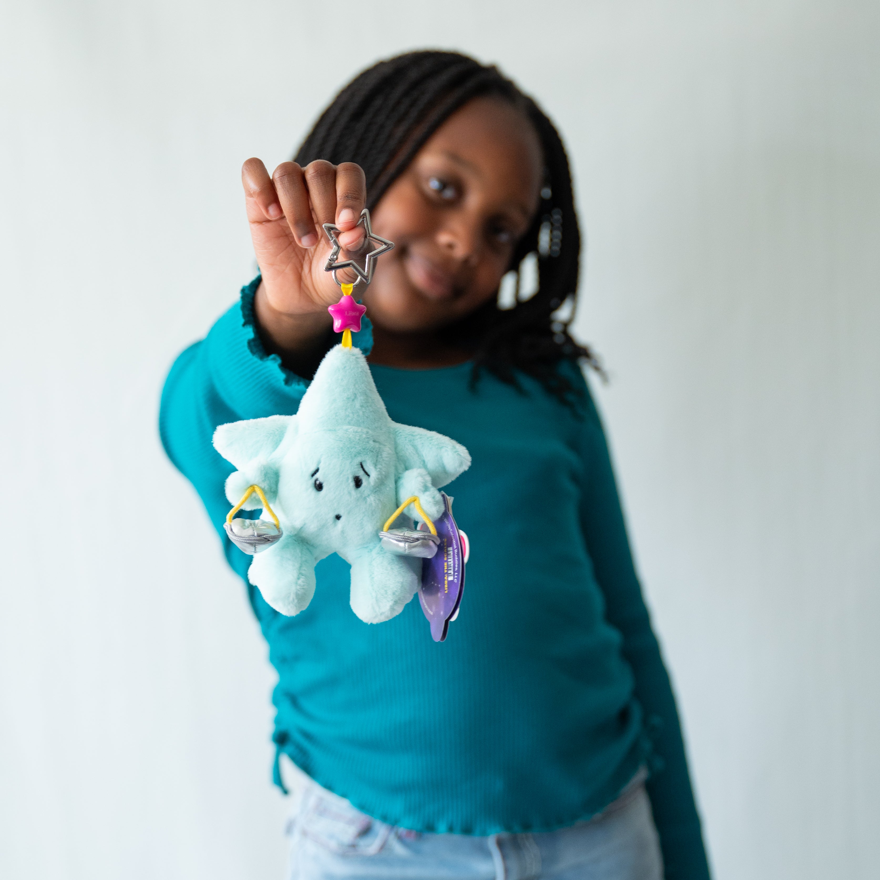 A young girl with braided hair wearing a teal long-sleeve shirt holds up a plush star-shaped Libra Zodiac keychain against a plain white background.
