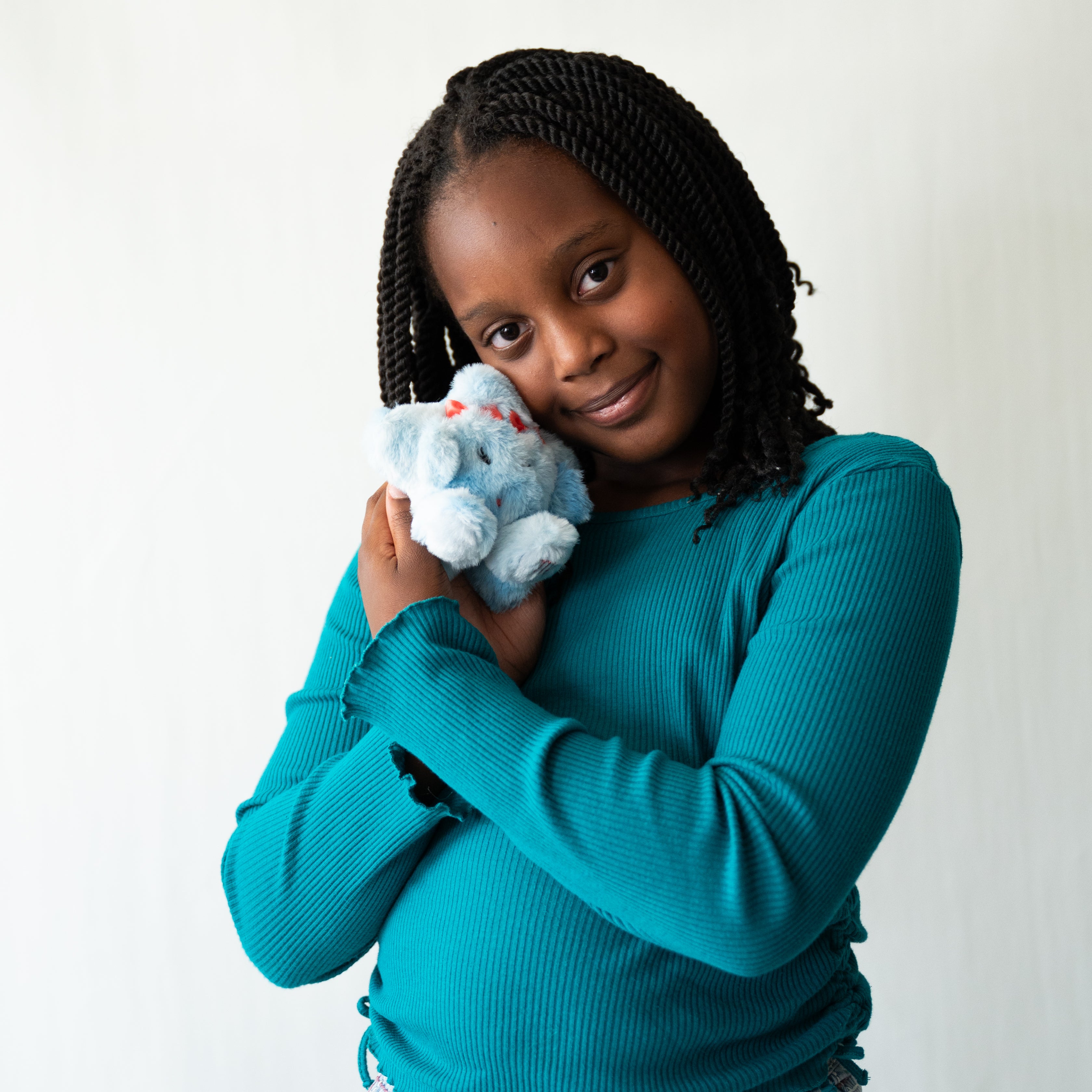 A woman with black braids holds a small light blue plush keychain against her chest, wearing a teal ribbed long-sleeve top, photographed against a plain light background.