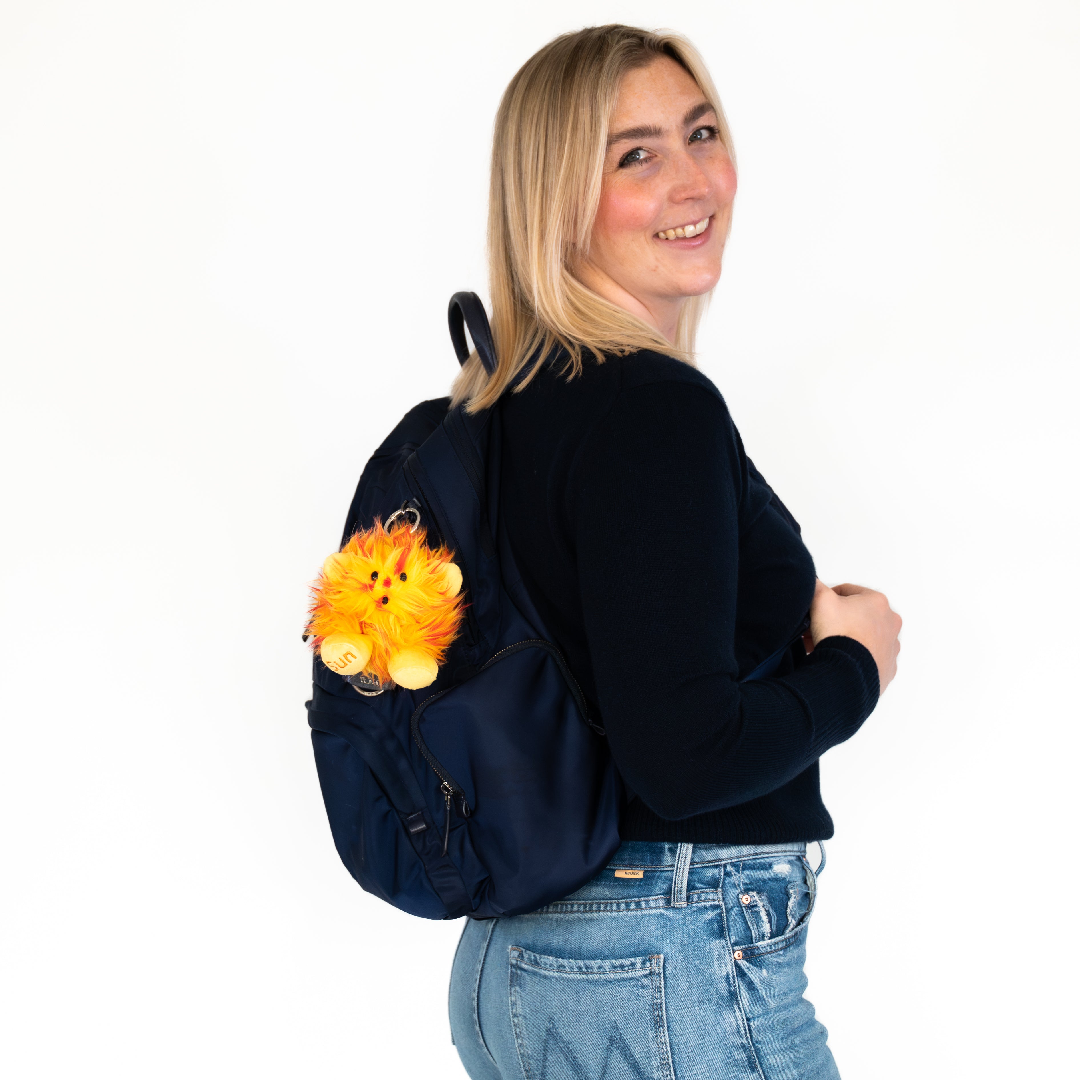 A woman wearing a navy blue backpack with a fluffy orange plush Key chain (Sun Nano) attached to the side, posing against a solid white background.