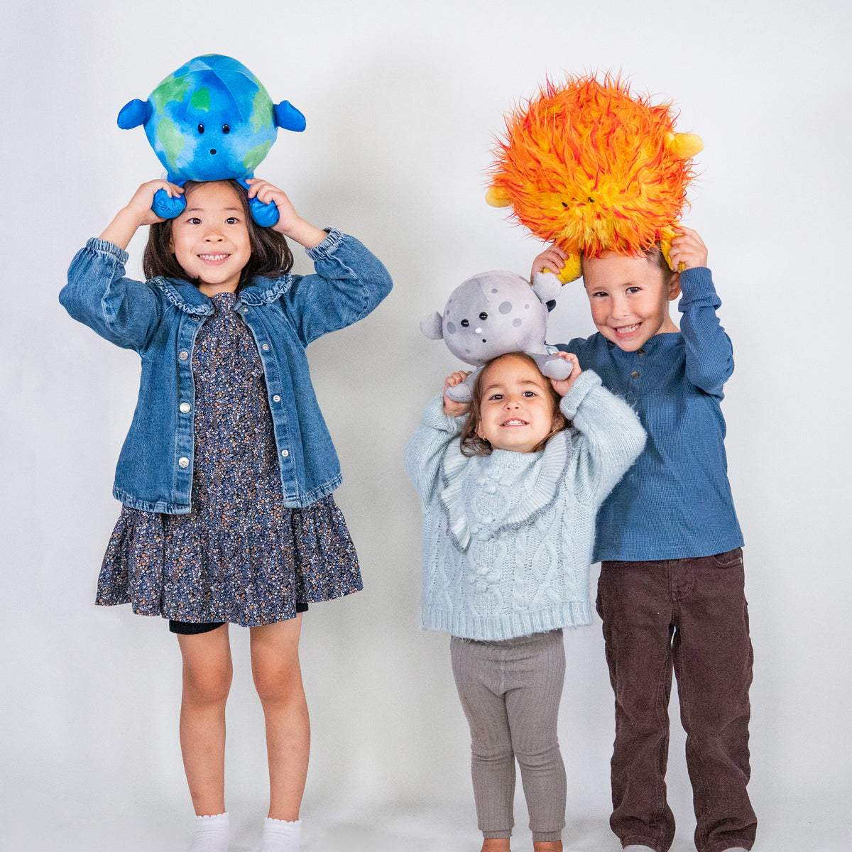 Three children each holding a Buddie from the Best Seller Bundle over their head, smiling and enjoying the product. Space plushies. Earth plushie. Moon Plushie. Sun plushie