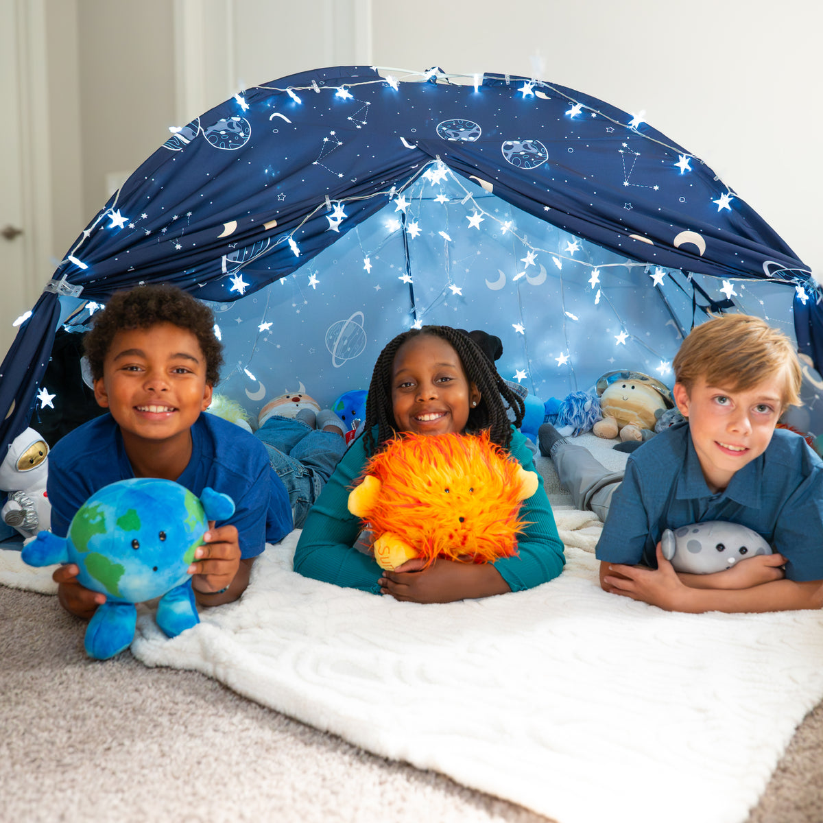 Three kids resting on their elbows holding space plushies are playing in a space-themed tent with the Best Seller Bundle. Earth Buddy, Sun Buddy, Moon Buddy.