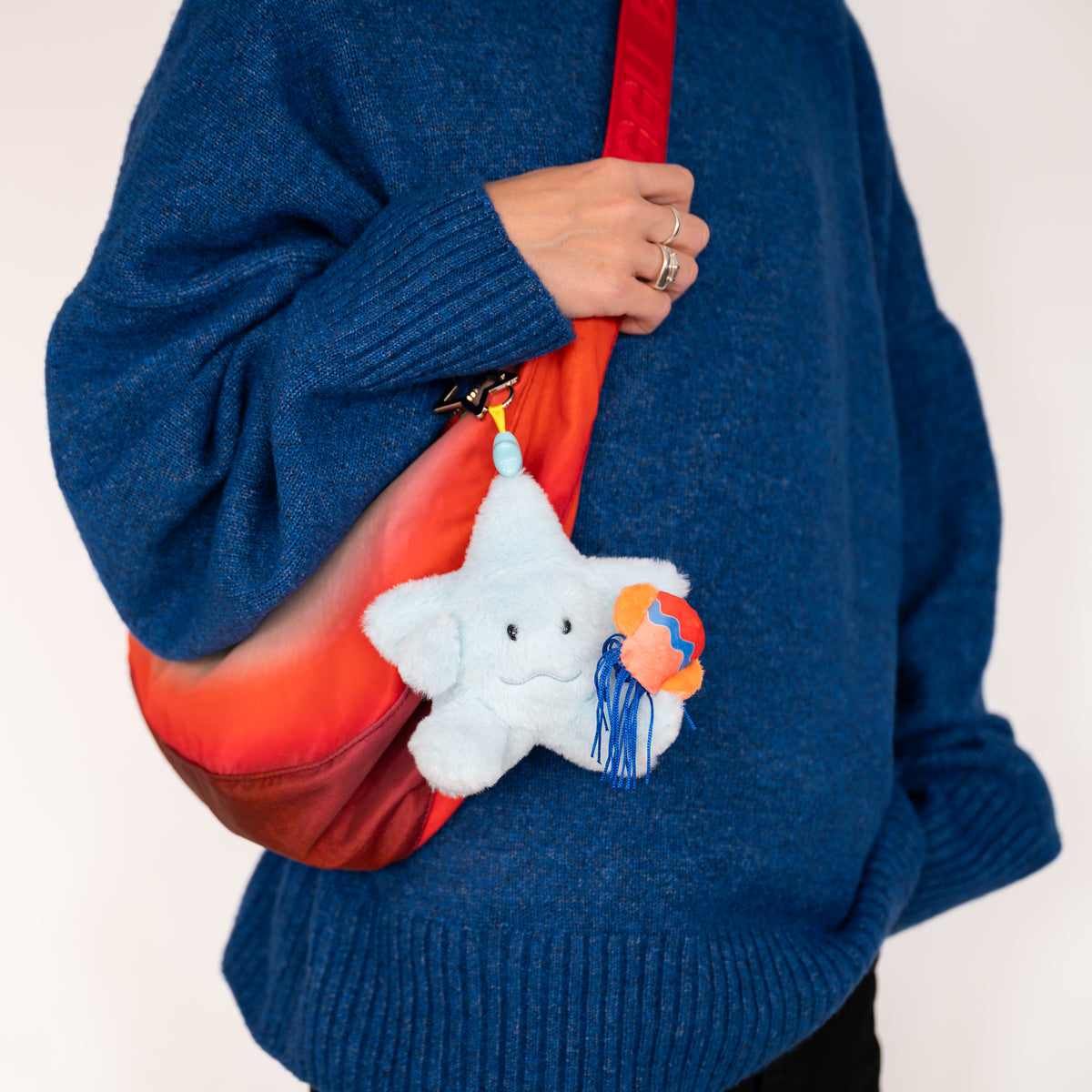 A person wearing a blue knit sweater carries a red and orange gradient shoulder bag with a white plush star charm holding a small colorful globe.