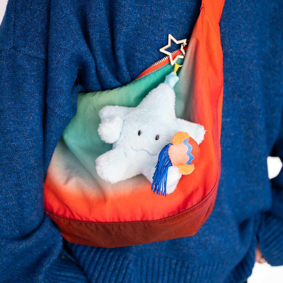A close-up of a person wearing a blue knit sweater and carrying a rainbow gradient shoulder bag with a white plush star toy and a small colorful tassel charm.
