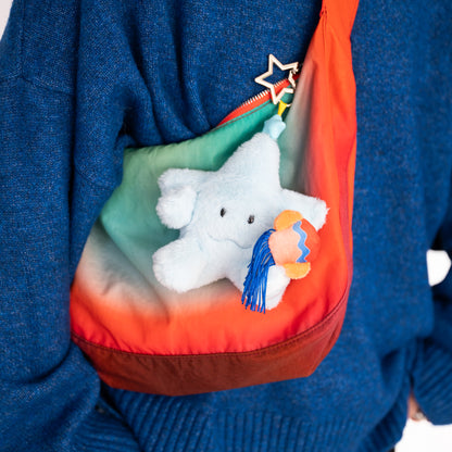A close-up of a person wearing a blue knit sweater and carrying a rainbow gradient shoulder bag with a white plush star toy and a small colorful tassel charm.
