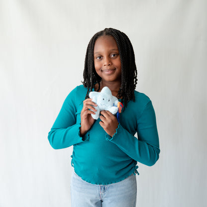 A young girl with black braids is smiling and holding a light blue plush star toy. She is wearing a teal long-sleeve top and light blue jeans, standing against a plain white studio background.