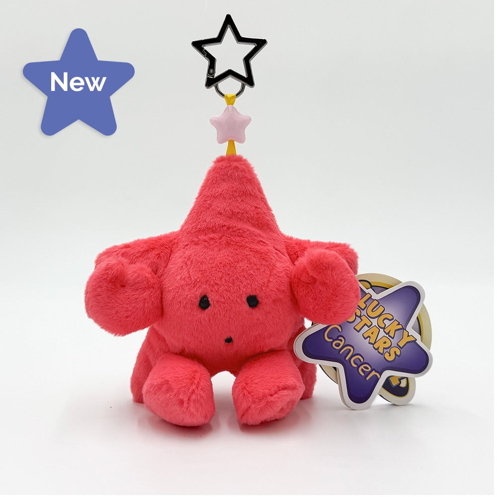 A pink fuzzy plush star keychain with a pointed top, black embroidered eyes and nose, a small light pink star, and a black star outline hanging above it. A purple and silver tag reading 'LUCKY STARS Cancer' is attached.