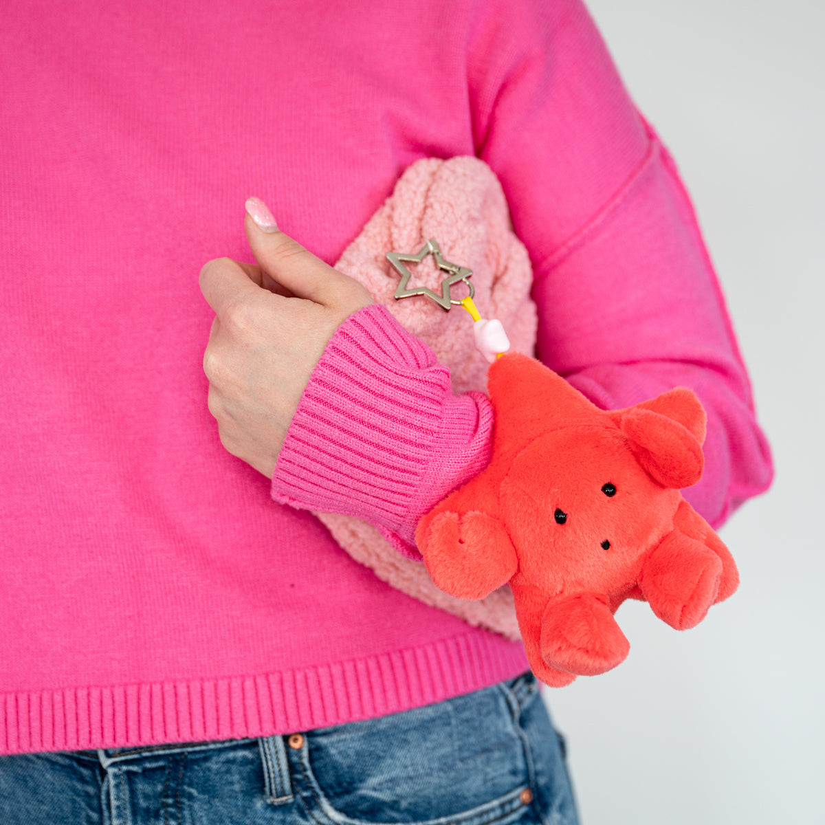 A woman wearing a bright pink sweater holds a pink fuzzy pouch on her arm. Attached to the pouch is a small plush star-shaped keychain in coral red, with a metal star keychain hanging from it.