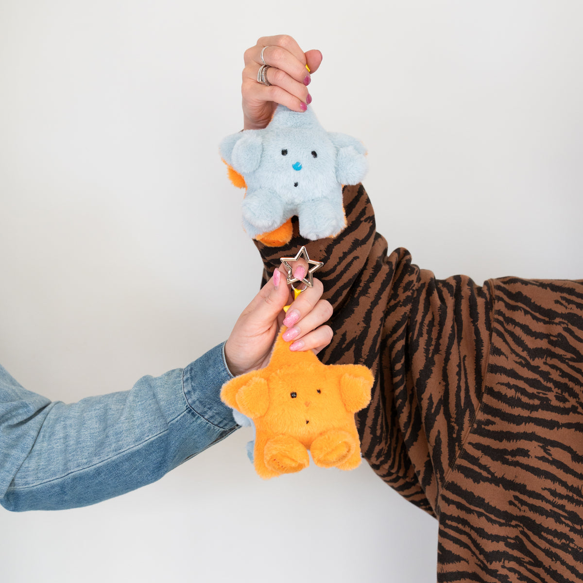 Two hands holding plush keychains against a plain white background. One hand holds a light blue star shape plush with a small orange star charm, while the other hand holds an orange star shape plush.