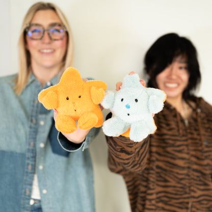 Two smiling people holding plush keychains in a bright indoor setting. The person on the left holds an orange star-shaped plush, and the person on the right holds a light blue star shaped plush.