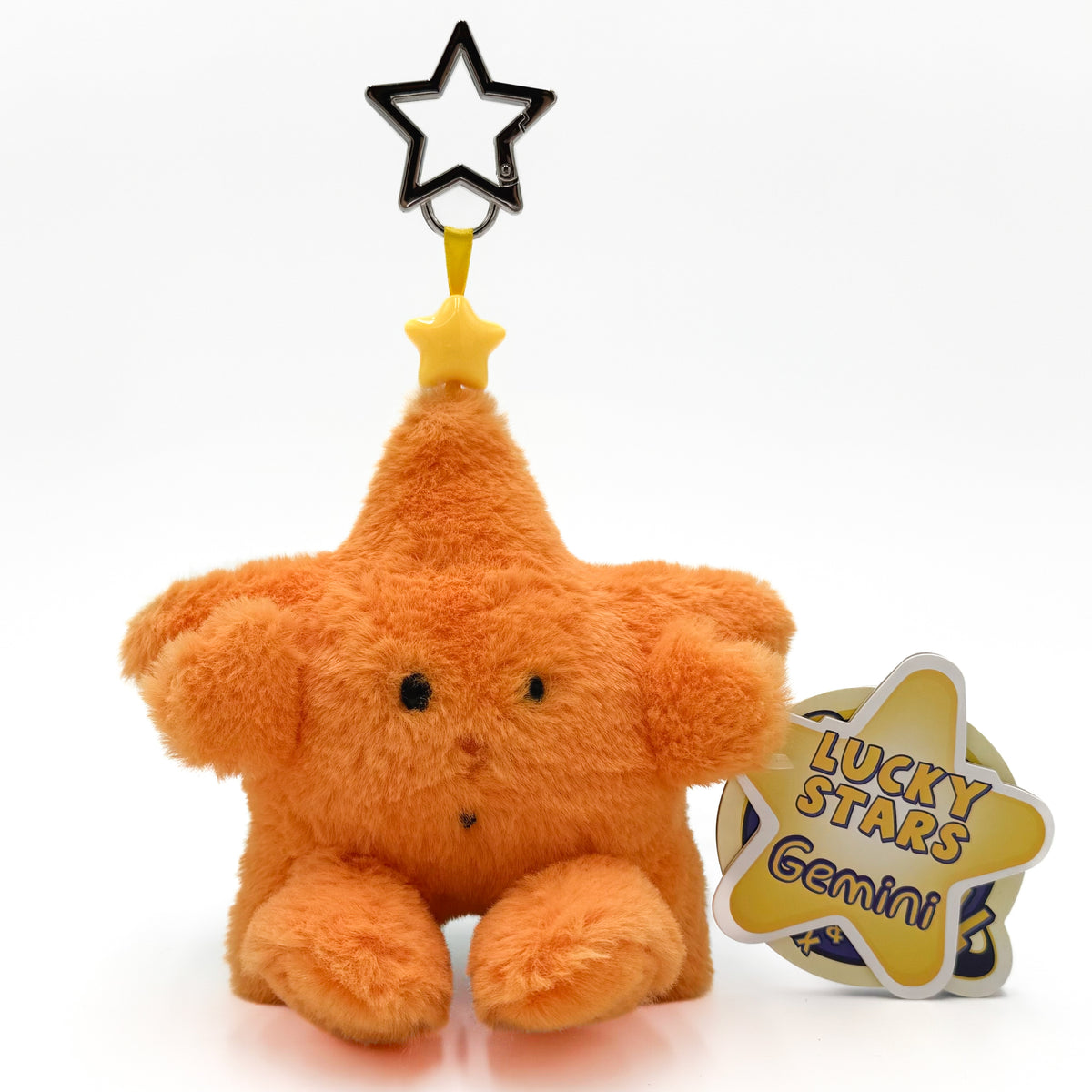 An orange fuzzy plush star keuchain with a pointed top and a black star outline hanging from a yellow loop, shown on a white background next to a yellow tag that reads 'LUCKY STARS Gemini'.
