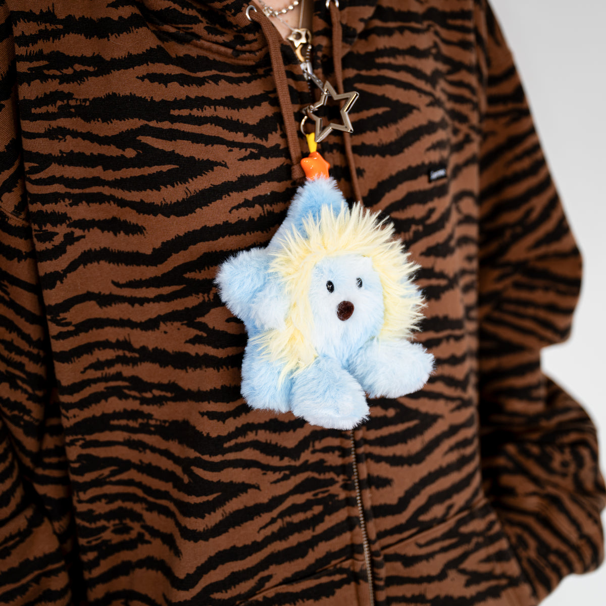 A plush star-shaped keychain or bag charm with a light blue body and fluffy cream-yellow mane, hanging from a brown zipper on a brown tiger-print hoodie.