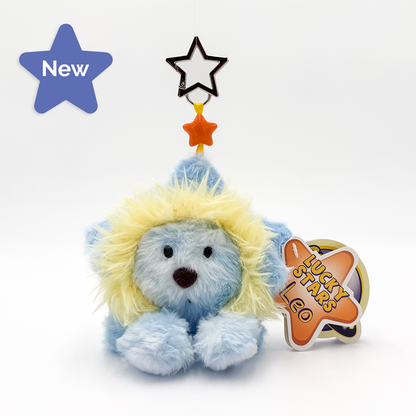 A plush lion toy in light blue and white with a fluffy yellow mane, sitting on a white background. It has a hanging star ornament above its head and a paper tag that reads 'LUCKY STARS LEO'.