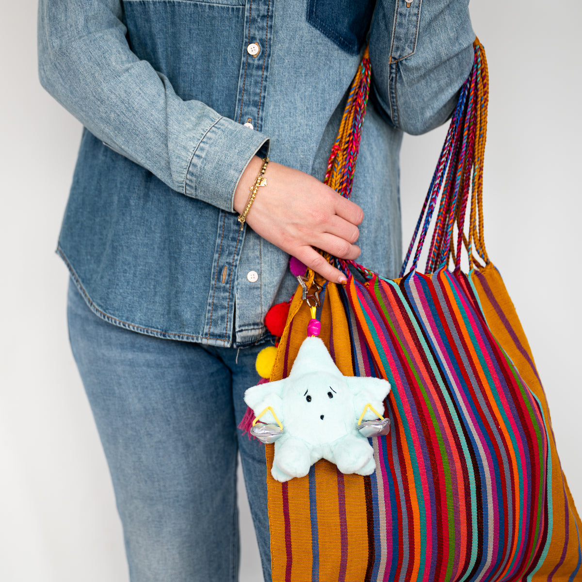 A person wearing a denim shirt and jeans holds a large striped tote bag with a light blue plush star keychain hanging from it. The bag has vertical stripes in orange, red, blue, yellow, and purple with braided shoulder straps.
