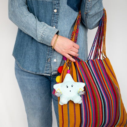 A person wearing a denim shirt and jeans holds a large striped tote bag with a light blue plush star keychain hanging from it. The bag has vertical stripes in orange, red, blue, yellow, and purple with braided shoulder straps.