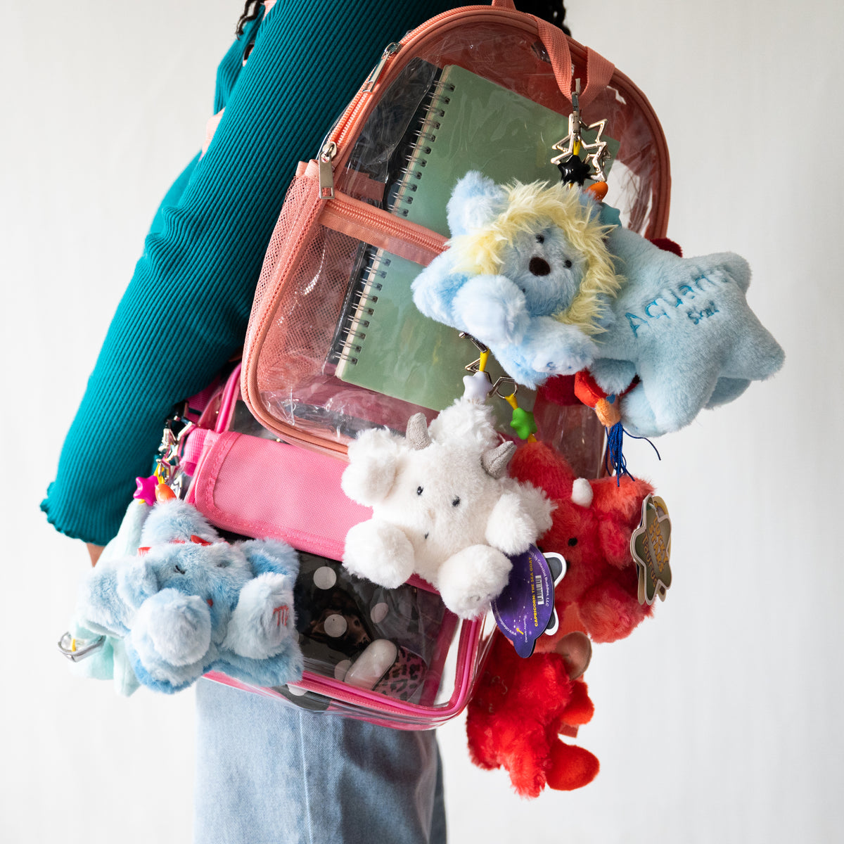 A person holds a clear backpack with a pink interior, filled with stationery and surrounded by several plush stuffed animals in blue, white, and red, against a plain light background.