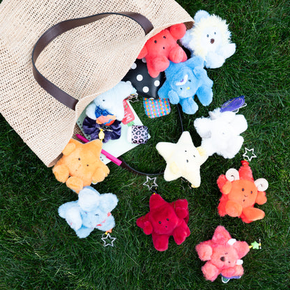A natural jute tote bag lies on green grass with several colorful plush star-shaped accessories spilling out of it. The stars are in assorted colors including red, blue, white, yellow, orange, and pink, and one has a small bow accessory attached.