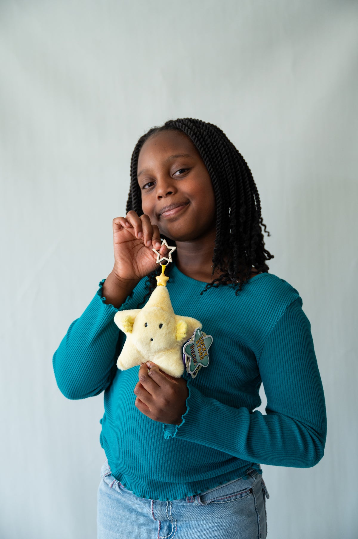 A young girl with braided hair wearing a teal ribbed long-sleeve top and light blue jeans, holding a yellow plush keychain star  with a silver star topper against a light gray studio background.