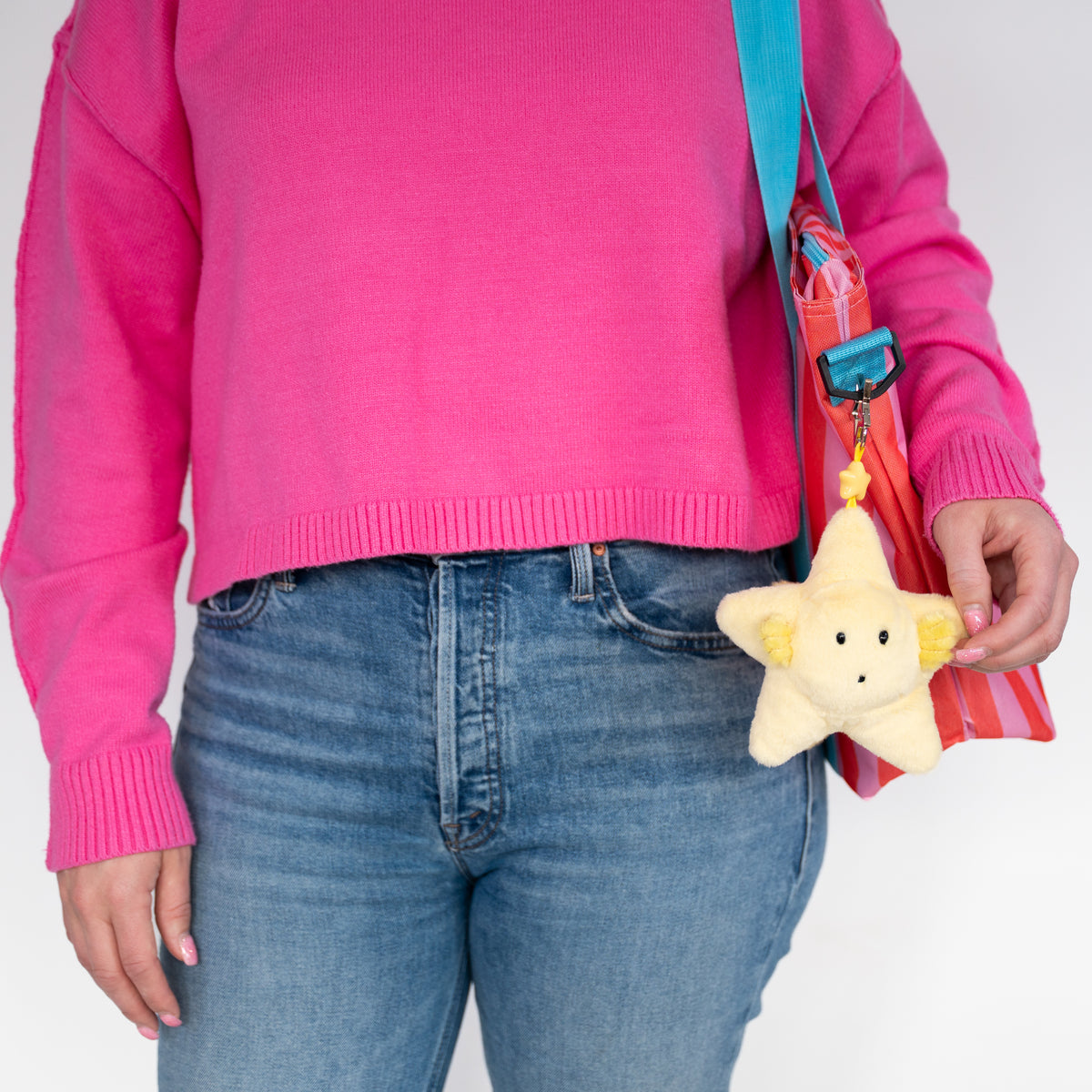 A person wearing a bright pink sweater and blue jeans holds a small yellow plush star keychain with a black face and yellow ears. A red and orange striped shoulder bag with a blue strap is also visible against a plain white background.