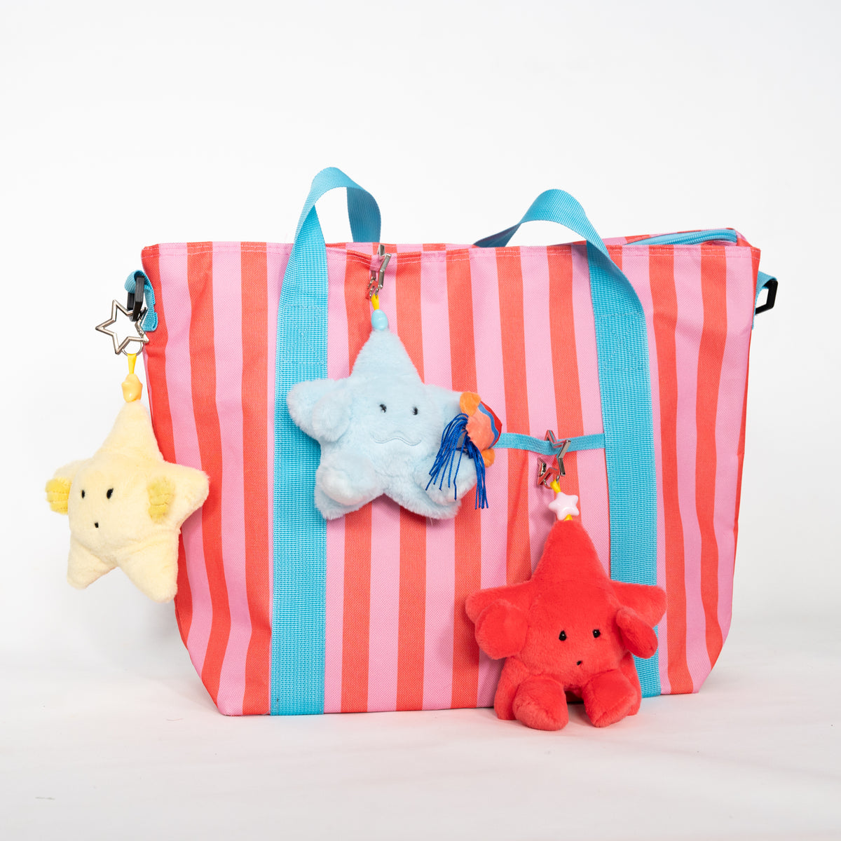 A striped tote bag with pink, coral, and turquoise vertical stripes, decorated with three plush star-shaped keychains in yellow, light blue, and red, hanging from metal rings on the side handles.