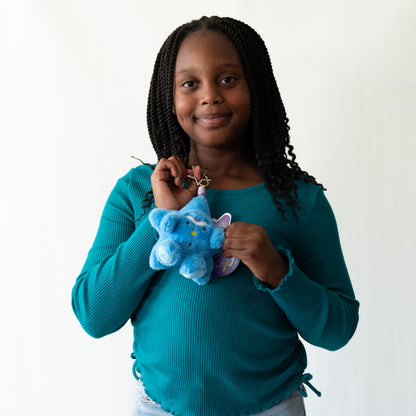 A young girl with long black braids smiles while holding a small blue plush star shaped keychain with a gold keyring, wearing a teal ribbed long-sleeve top against a plain white background.