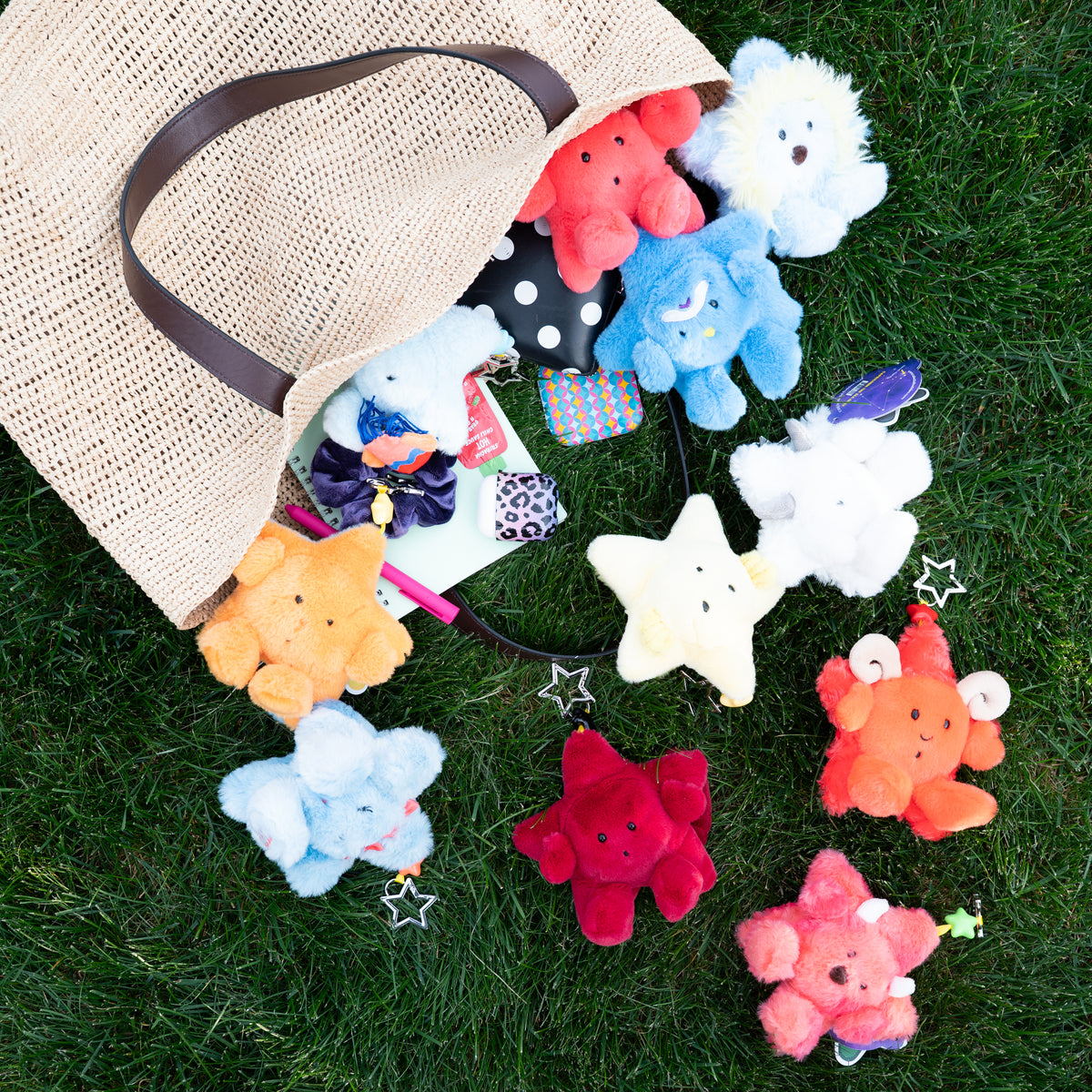 A natural jute tote bag lies on green grass with several colorful plush star-shaped keychains spilling out of it. The stars are in assorted colors including red, blue, white, yellow, orange, and pink, and one has a small bow accessory attached.