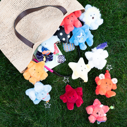 A natural jute tote bag lies on green grass with several colorful plush star-shaped keychains spilling out of it. The stars are in assorted colors including red, blue, white, yellow, orange, and pink, and one has a small bow accessory attached.