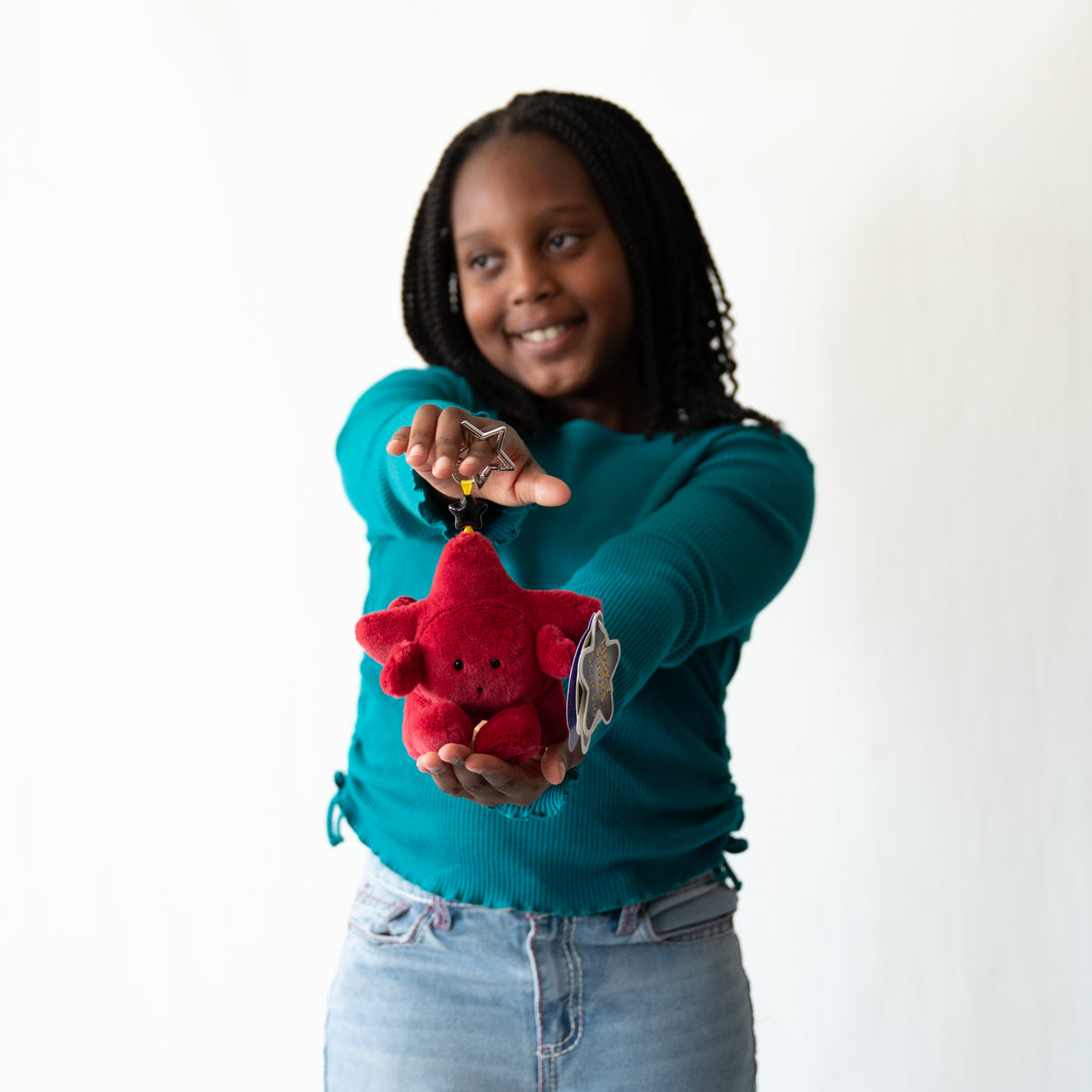A young girl with braided hair wearing a teal long-sleeve shirt and light blue jeans holds out a small red plush star keychain to the camera against a plain white background.