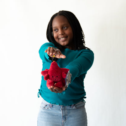 A young girl with braided hair wearing a teal long-sleeve shirt and light blue jeans holds out a small red plush star keychain to the camera against a plain white background.