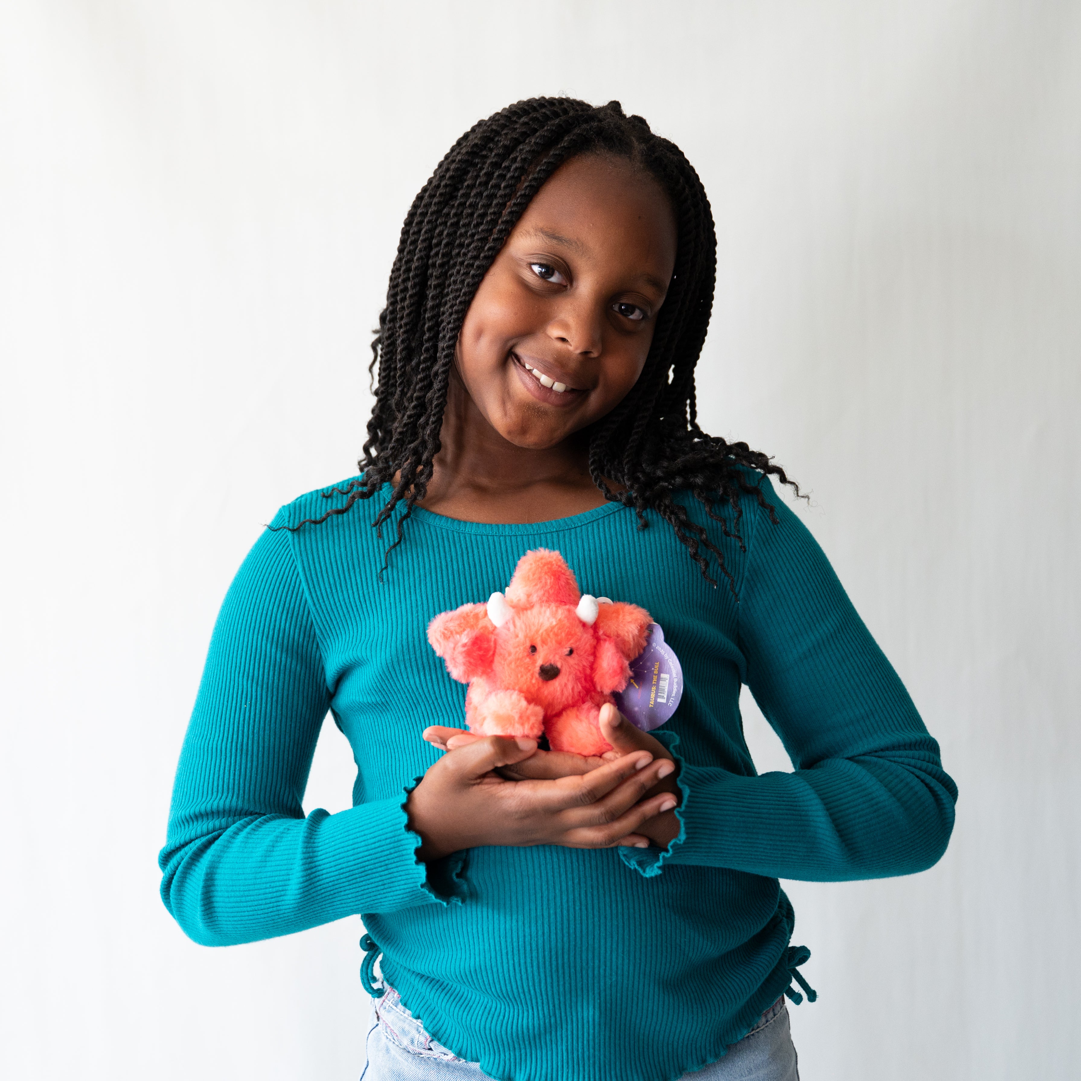 A young girl with braided hair smiles while holding a small pink keychain with a purple tag, wearing a teal long-sleeve top against a plain light background.