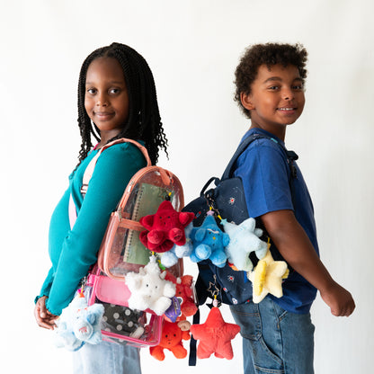 A young boy and girl standing back-to-back wearing backpacks decorated with colorful plush star keychains photographed against a plain white background.