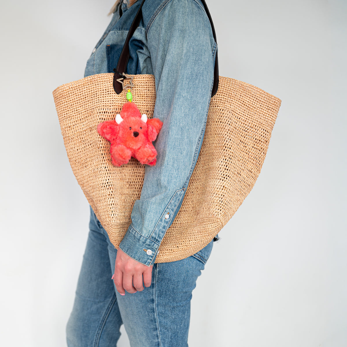 A woman wearing a denim jacket and jeans carries a large woven straw tote bag with a black shoulder strap. A small red plush keychain charm hangs from the bag.