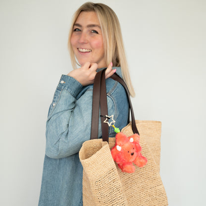 A woman in a denim shirt carries a large woven straw tote bag with dark brown leather handles. The bag is decorated with a small star shaped keychain and a silver star charm, set against a plain light gray background.