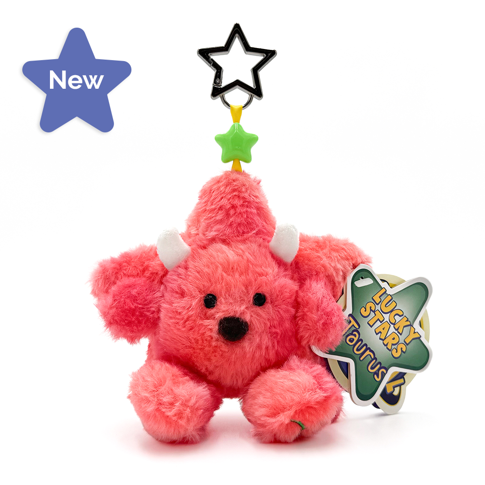 A pink plush keychain with small white horns and a black nose, attached to a green star-shaped tag that reads 'Lucky Stars Taurus.' It also has a black star and green star charm on a yellow string above it, photographed on a clean white background.
