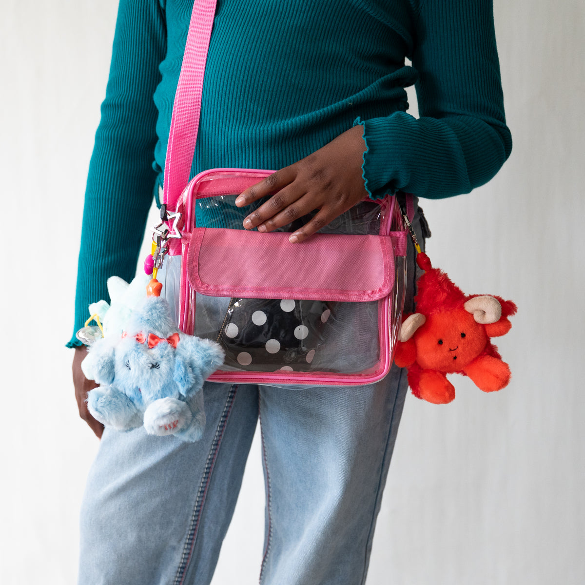 A person wearing a teal ribbed long-sleeve top and light blue jeans carries a pink and clear crossbody bag with a bright pink strap. The bag has a clear front pocket showing a black-and-white polka dot item inside, and three plush keychains hang from the side: two light blue ones and a bright orange-red one.