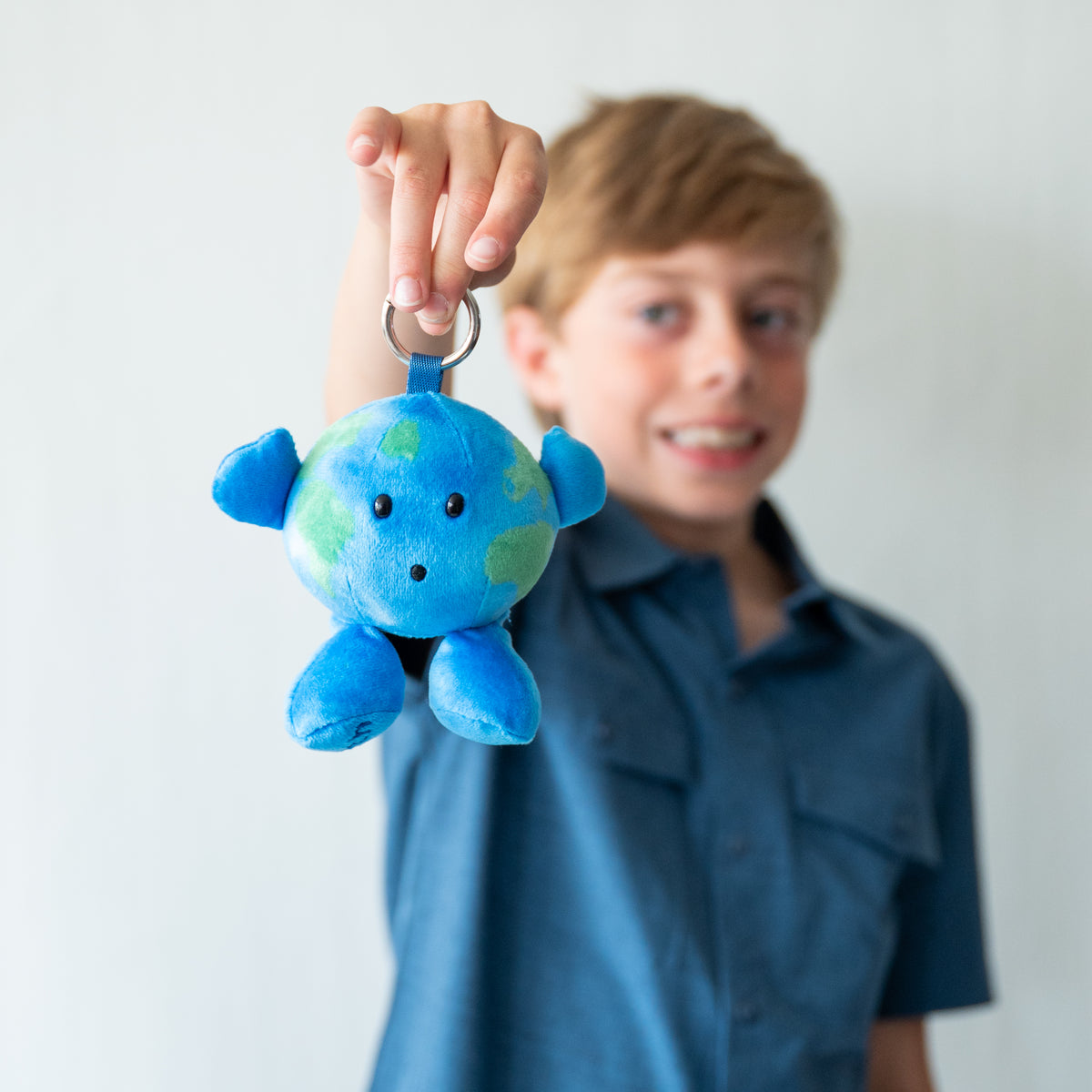 A young boy in a blue shirt holds up a small blue and green plush Earth Nano  keychain with a metal key ring, with his face blurred in the background against a light gray wall.