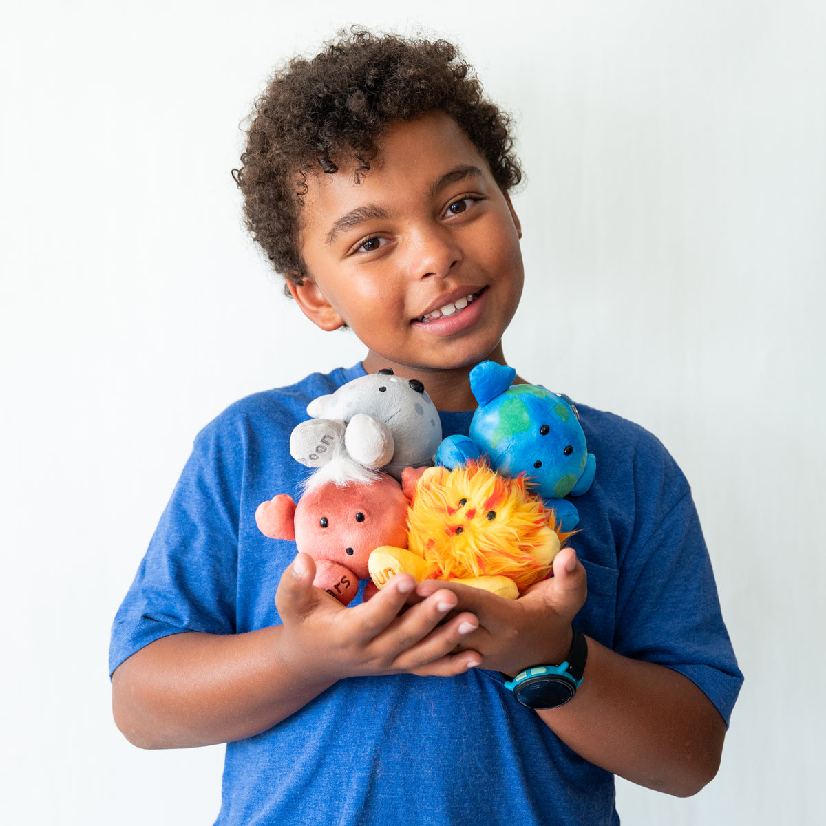 A smiling young boy with curly dark hair holds a small pile of colorful plush animal toys against a plain white background. He is wearing a blue heathered t-shirt and a black smartwatch.