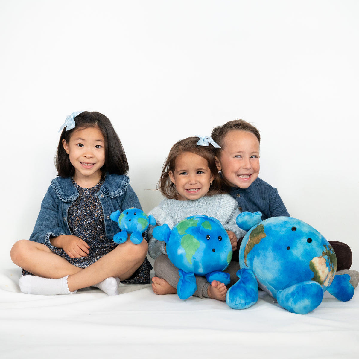 Three children sitting together on a white floor against a white wall, each holding a blue plush Earth toy. The children are wearing blue outfits and bow headbands, and the plush toys are bright blue with green landmasses.