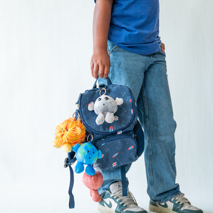 A child wearing a blue t-shirt and jeans holds a small blue astronaut backpack with attached plush toys against a plain white background.