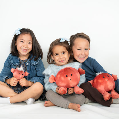 Three children sitting together on a white floor against a white wall, each holding a plush toy. The toys are round and coral-red with small black embroidered eyes and mouths and short flippers or tentacle-like limbs.