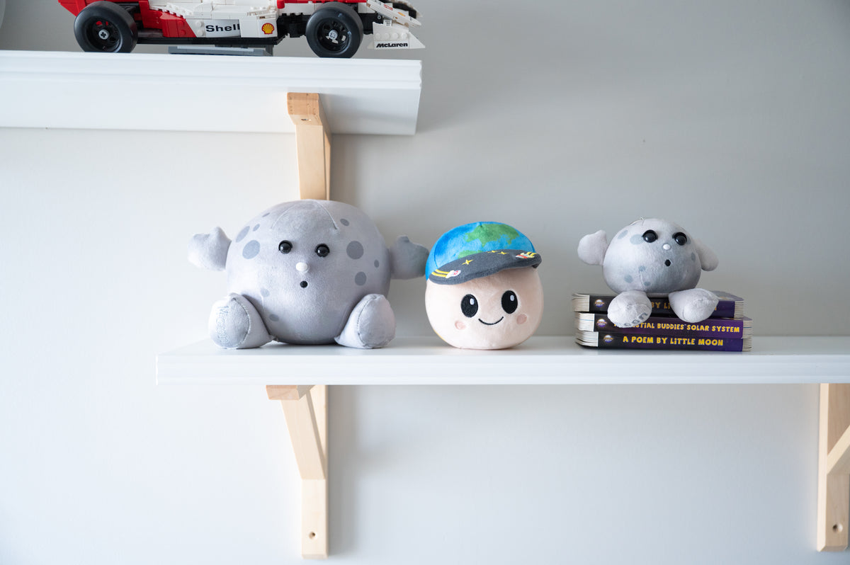 Three cute plush toys shaped like planets (Moon Buddy, Rise Buddy and Nano Moon ) displayed on white shelves against a light gray wall. 