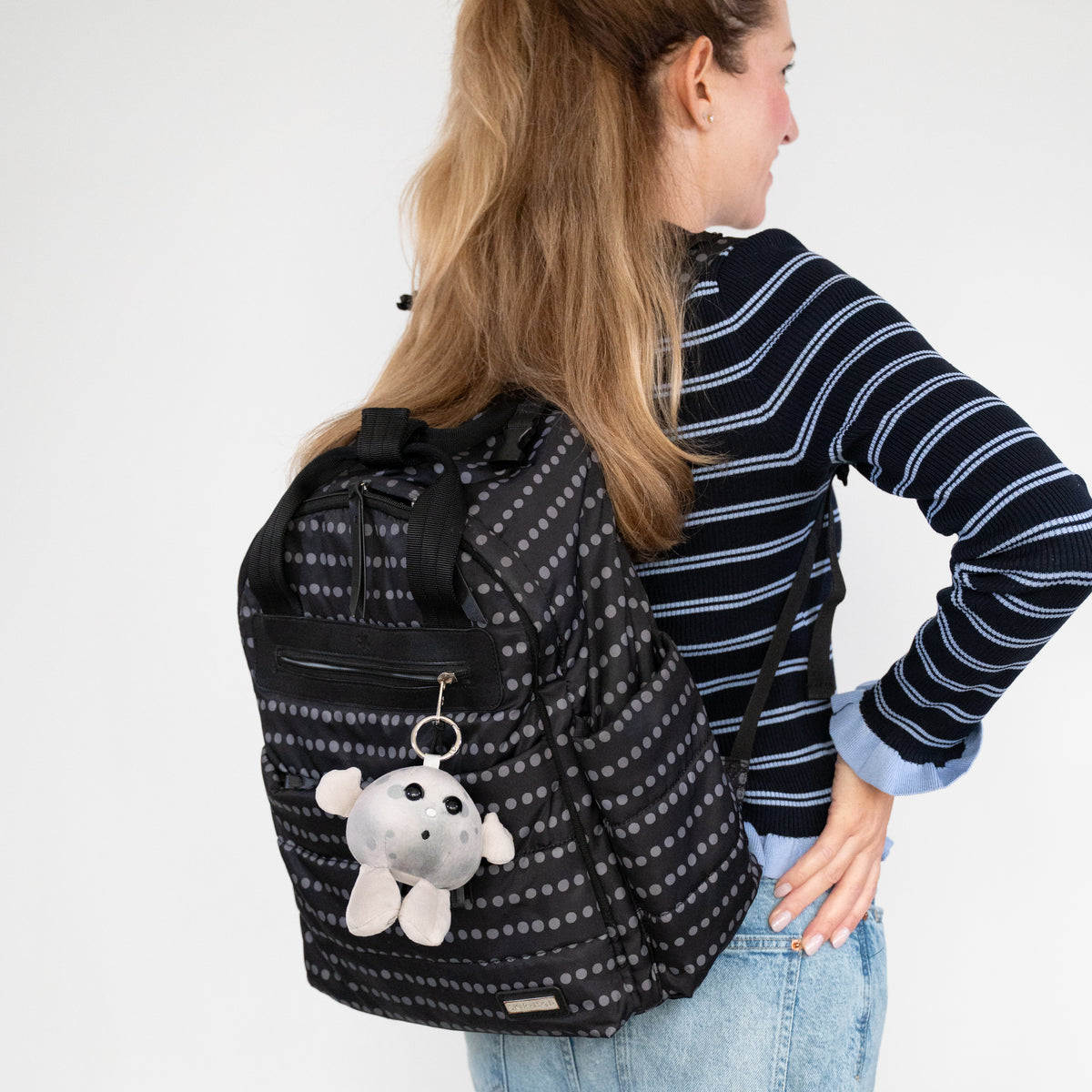 A woman wearing a navy and white striped sweater and blue jeans is shown from the back, carrying a black backpack with a gray polka dot pattern and Nano Moon plush keychain.