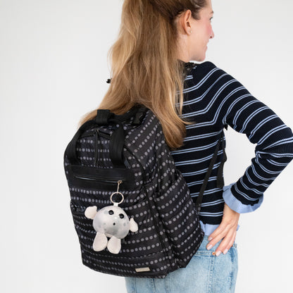 A woman wearing a navy and white striped sweater and blue jeans is shown from the back, carrying a black backpack with a gray polka dot pattern and Nano Moon plush keychain.