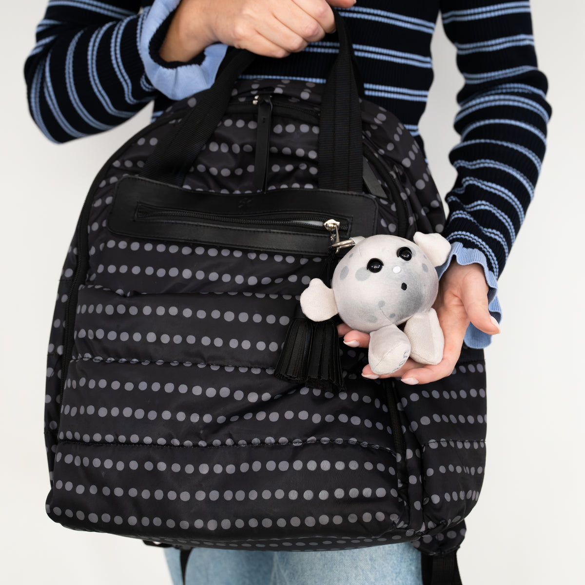 A person holds a black puffer-style tote bag with a gray polka dot pattern. The bag has black handles, a front zip pocket, and a gray plush character charm with a black tassel.