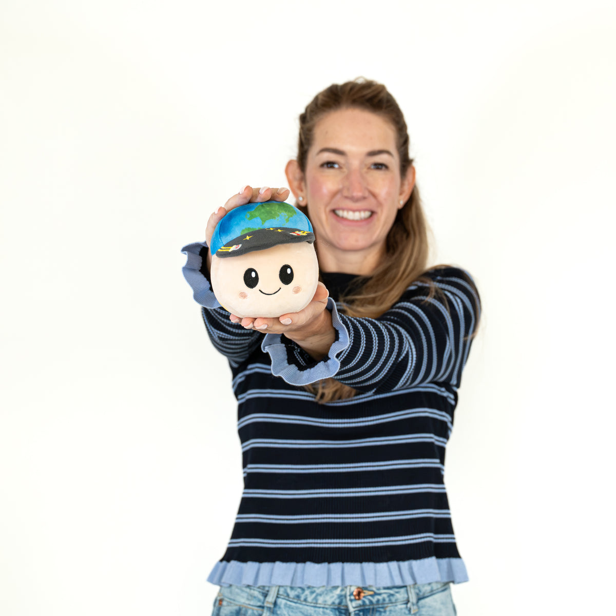 A smiling woman wearing a navy and light blue striped long-sleeve top holds up a small plush toy with a cartoon face and a blue-and-green globe cap against a plain white background.