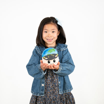 A young girl with dark hair wearing a blue denim jacket and a dark floral dress holds a small plush toy shaped like the Earth in her hands against a plain white background.