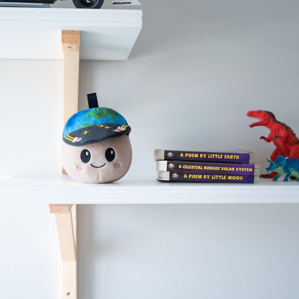 A cute, round beige plush toy with a smiling face and a colorful globe-patterned cap, sitting on a white shelf next to books and a red dinosaur toy.